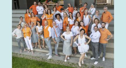 Eine Gruppe Menschen in orange, weiß, grau und silber gekleidet, steht auf einer Treppe und lacht in die Kamera.